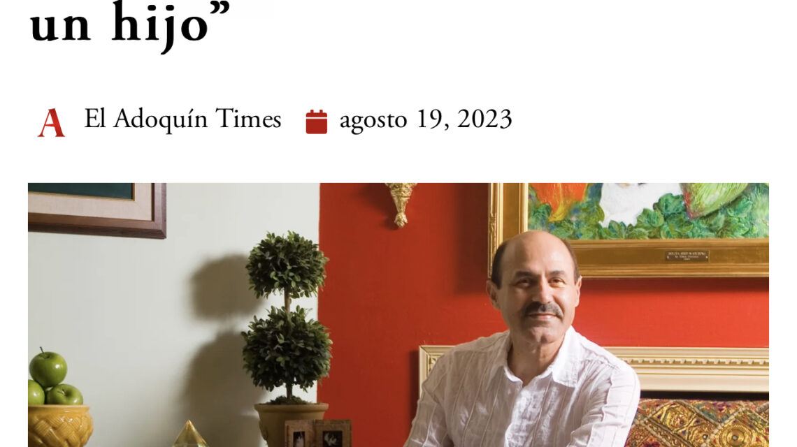 A man with a mustache in a white linen shirt and jeans sits with his legs crossed on a bench with an embroidered pillow in front of a red wall
