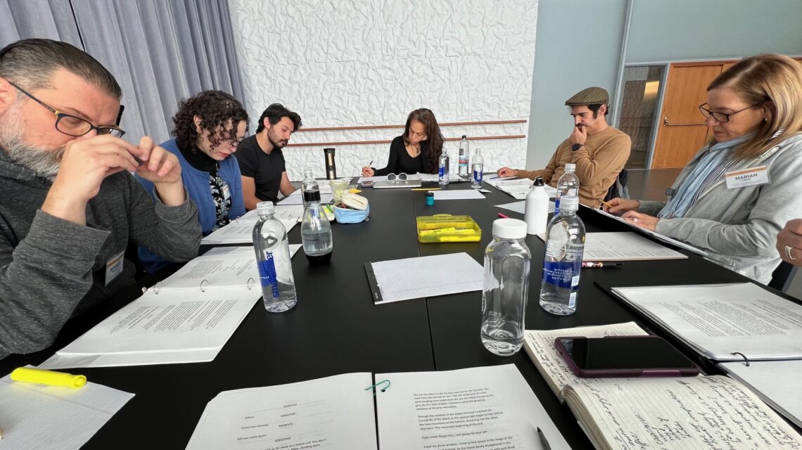 A group of six men and women sit around a large table looking at scripts in 3-ring binders