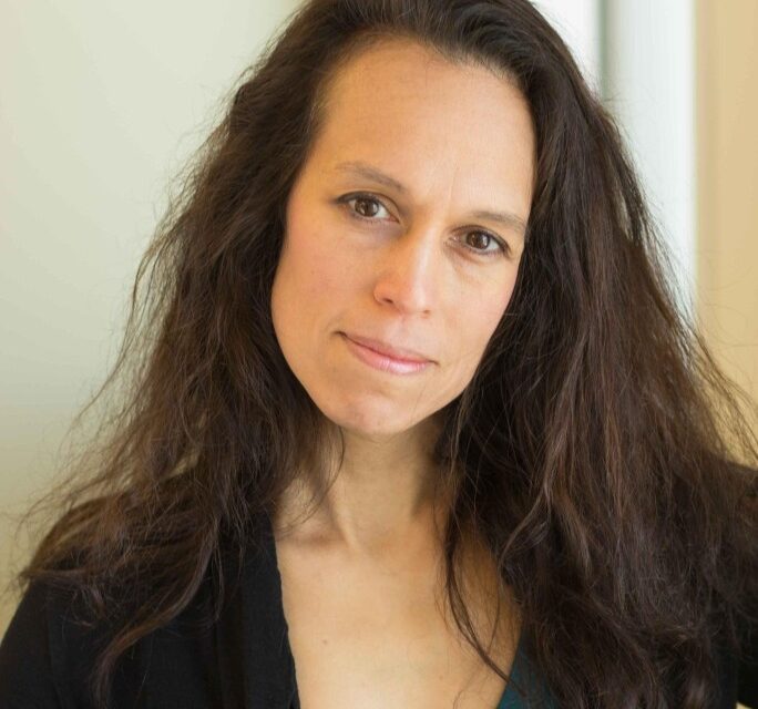 A woman with long brown hair and brown eyes wearing a dark v-neck shirt tilts her head and smiles