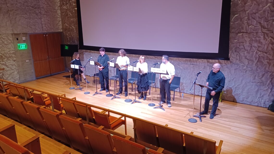 A row of 5 men and women stand on a stage in front of music stands labeled with character names as a man on the far right dressed in black reads from a script
