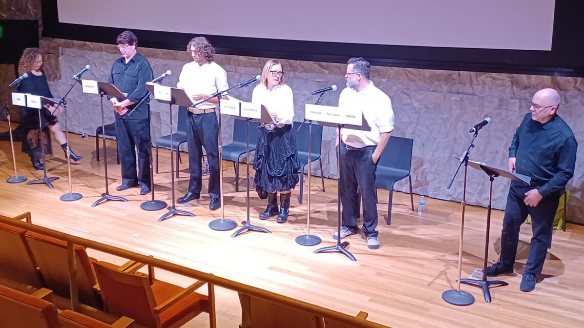 A row of 5 men and women stand on a stage in front of music stands labeled with character names as a man on the far right dressed in black reads from a script