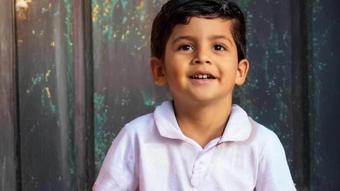 A young boy with brown hair and eyes sits on a stoop in a white polo shirt